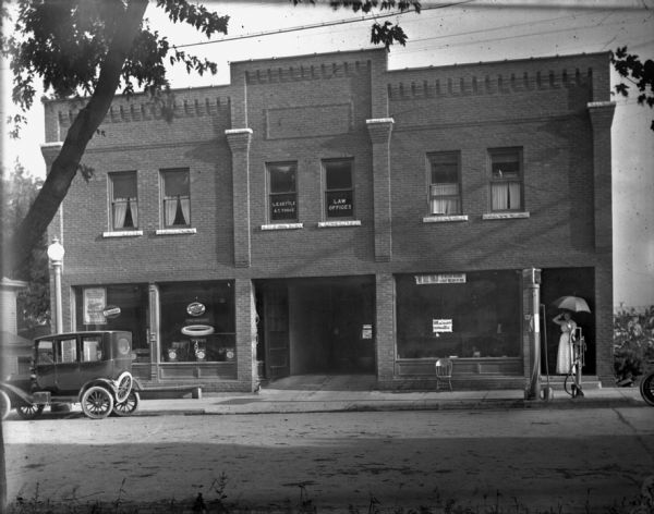 View across street towards car dealership on the first floor of a two-story brick building. The first Louis Schoelkopf car dealership was on State Street, and later moved to Pinckney Street. A car is parked at the curb on the left in front of the large front display windows. There is a woman in a long summer dress with an umbrella standing in the open foyer of a doorway on the right, and in front of the her is a gas pump and an air pump on the sidewalk at the curb.

The second floor has law offices for L.E. Gettle and A.T. Torge on one side, and the office of Dr. O. Ishmael on the other.