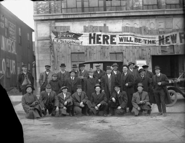 View of group of men, and a young boy, gathered for a group portrait in front of the 210 E. Washington Avenue dealership. The Schoelkopf name has been added to the front entrance and the banner moved down to cover the as yet unfinished entrance. The building on the left has a sign painted on the exterior that reads: "Universal Flour."