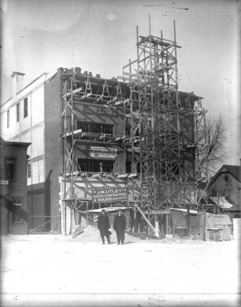 Another view of the Schoelkopf construction site at 210 E. Washington Avenue. Two men stand in the street in front, one of whom may be Louis Schoelkopf. The J.W. Utley Co. General Contractors and Builders sign is behind the two men on the scaffolding, and a banner on the brick building reads, in part: "This Bldg is Heated by Kewaunee Fire Box Boiler."