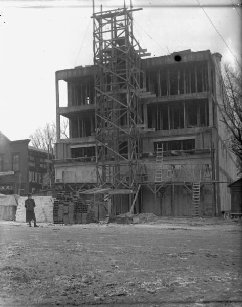View across street towards the construction at 210 E. Washington Avenue. There is scaffolding in front of the building, and Emma Schoelkopf is standing in front wearing a fur wrap and holding a muff. There are stacks of bricks and other building materials in front of the main entrance.

The Universal Flour building next door also houses a taxi and a printing business.