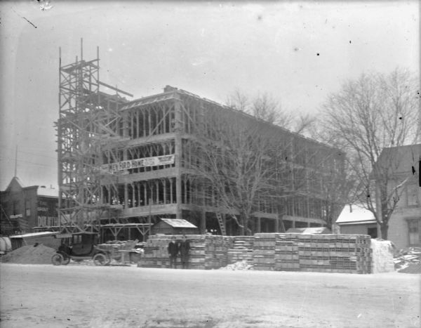 View across street towards two men standing in front of the Schoelkopf dealership at 210 E. Washington Avenue as construction progresses on the three-story building. A banner on the building reads: "New Ford Home." A Ford car is parked on the street, and building materials are stacked along the side of the street. Next door is the Duluth Universal Flour and Baking Company.