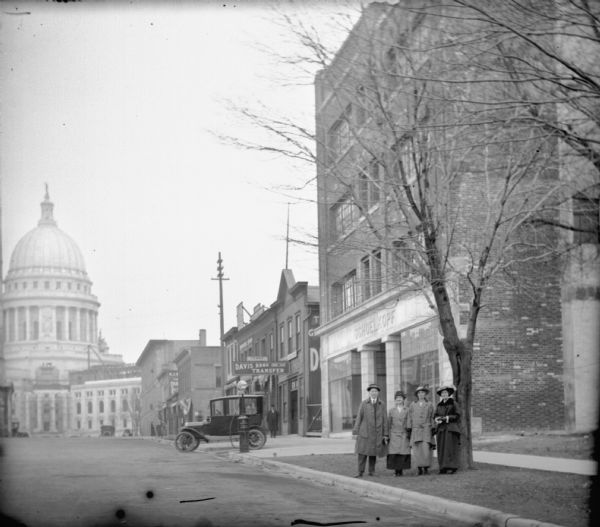View up E. Washington Avenue towards a group of four smiling people standing on the curb near the Schoelkopf Automobile dealership soon after construction was completed on the building. Emma Schoelkopf wears a checked coat, and the other people are unidentified. The Wisconsin State Capitol is in the background.