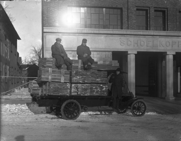 View from street of three men posing on a Ford truck loaded with wood boards. The truck is parked in front of the Schoelkopf Ford dealership at 210 E. Washington Avenue. Snow is on the ground. The men are dressed in winter hats and coats.