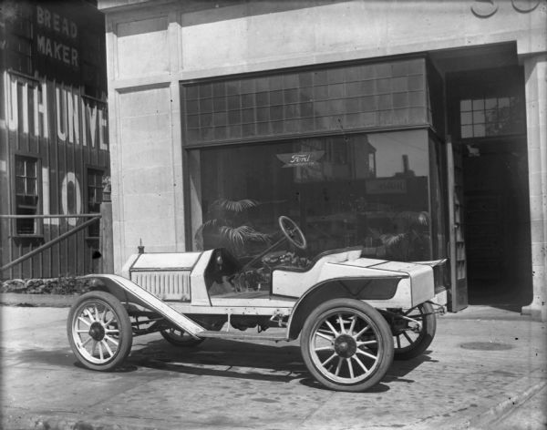 Light-colored Ford speedster is parked on the sidewalk in front of Schoelkopf's dealership at 210 E. Washington Avenue. There is a Ford sign in the show window. The building next door on the left has a sign painted on the wall for "Duluth Universal Flour."
