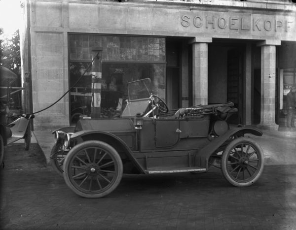 Small roadster parked in front of Schoelkopf's dealership at 210 E. Washington Avenue. A man stands near the entrance on the right. On the far left a car is attached to the air pump. The dealership has a Ford sign in the show window.