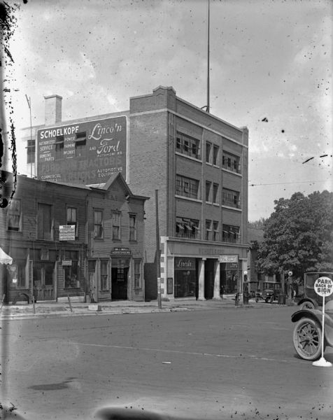 View from across street towards the Schoelkopf automobile dealership at 210 E. Washington Avenue. Above the show windows flanking the entrance are signs for "Ford" and "Lincoln." There is also a Ford sign on a pole outside the shop near air and gas pumps. A large sign painted on the side of the building near the roof advertises Ford and Lincoln automobiles, power farming implements, and other automotive supplies. The building next door at 208 has a horseshoeing business on the ground floor.