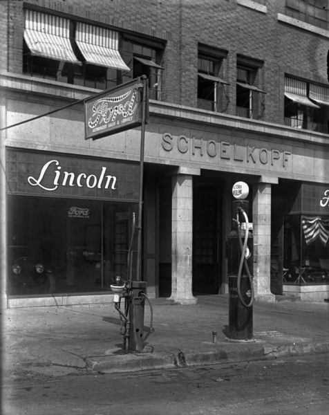View from street of the facade of the Schoelkopf Automobile dealership at 210 E. Washington Avenue. "Lincoln" has been added to the Ford dealership with a sign above the show window. There is a small Ford sign below it and also on a pole over the sidewalk. Underneath the Ford sign at the curb is an air pump and a water bubbler. A "Filtered Gasolene" pump is at the curb on the right.
