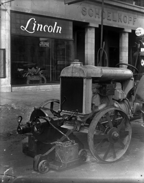 Fordson tractor parked in the street in front of Louis Schoelkopf's building at 210 E. Washington Avenue. The tractor has a battery on wheels attached to the front of it. There is a "Filtered Gasolene" pump on the curb behind the tractor.