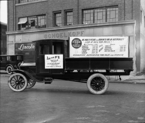 View from street towards a Ford truck parked in the street in front of the Schoelkopf Lincoln Ford Dealership at 210 E. Washington Avenue that is part of the New Trade Display Parade. Posted on the side of the truck are two signs. The one on the cab reads: "Ask for No 6, $603.58, Schoelkopf Authorized Ford Sales and Service." The sign over the truck bed reads, in part: "Henry Ford 'The Peoples Friend' Has Made It Possible for Everyone to Own an Automobile!" In the background on the left is an automobile with a spare tire mounted on the rear that reads: "The New Lincoln..."