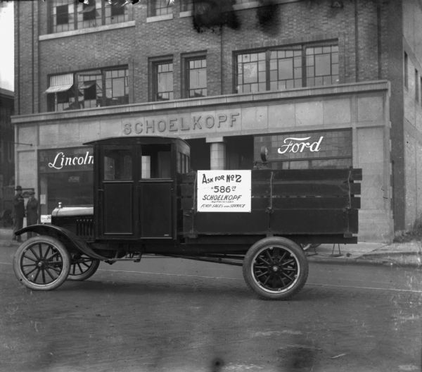 View from street towards a truck parked in front of the Schoelkopf Lincoln Ford Dealership at 210 E. Washington Avenue that is part of the New Trade Display Parade. Posted on the side of the truck is a sign that reads: "Ask for No 2 $586.07, Schoelkopf Authorized Ford Sales and Service." In the background on the left two men stand at the curb.