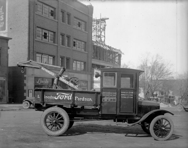View from street of the Lincoln-Ford Fordson tow truck, with "authorized service" printed on the tow truck arm. On the cab of the tow truck is a line drawing of the Schoelkopf building as it will look when expansion is completed. Behind the tow truck, the construction on the Schoelkopf building expansion at 210 to 216 E. Washington Avenue has reached the second floor addition.