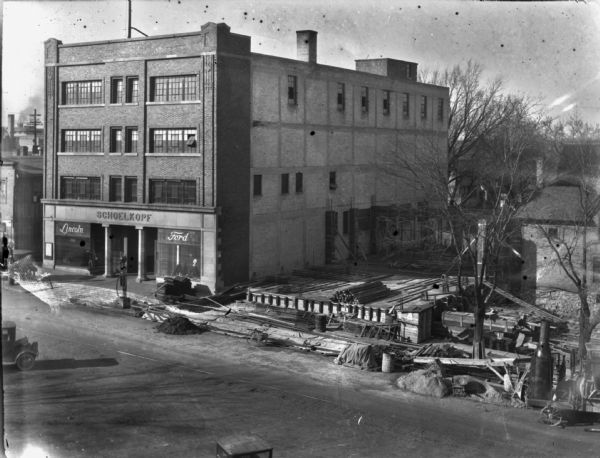 Elevated view of Lincoln Ford dealership at 210 E. Washington Avenue during its expansion to include the rest of the block to 216. Building materials and construction equipment are in the yard and along the curb. The two show windows flanking the entrance to the dealership have signs that advertise Lincoln and Ford.