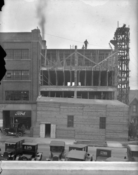 Elevated view from across street of the construction to expand the Schoelkopf building at 210-216 E. Washington Avenue. Men from the construction company are working on the third and fourth floor of the expansion. There are automobiles parked in the street in the foreground. On the sidewalk in front of the dealership is a Ford car.
