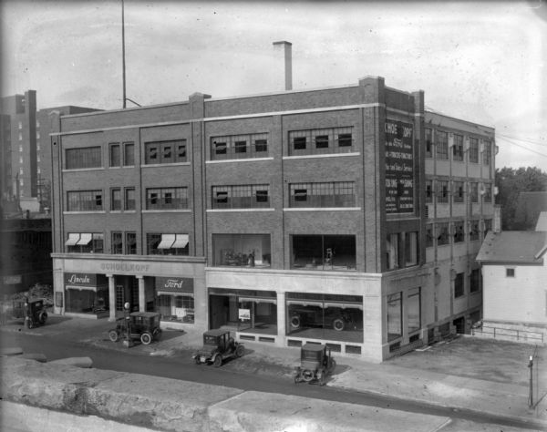 Elevated view from across street of 210-216 E. Washington Avenue. Four cars are parked on the sidewalk in front of the building. The expansion of the Schoelkopf Ford Dealership building is complete. In the left window of the original building, the small Ford sign under the Lincoln sign is no longer there. The Ford brand sign suspended from a pole on the street is still there. The new half of the building has signage on the first floor for "Cars, Trucks, Tractors." The second floor windows of the building show examples of Louis Schoelkopf's new business, the Air-Lec Automated Door Opener.