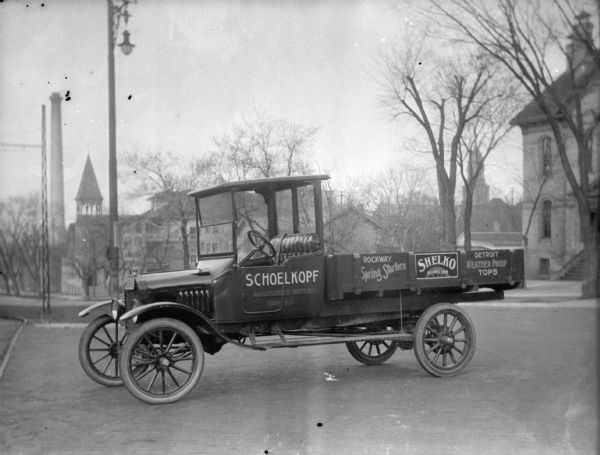 The Schoelkopf advertising truck parked on the street near the store at 210-216 E. Washington Avenue. The truck advertises a number of products: Rockway Spring Starters, Shelko Supplies and Detroit Weather Proof Tops. In the background down the hill is the steeple of St. Patrick Church on on E. Main Street, as well as a smokestack and an unidentified church steeple on E. Washington Avenue.