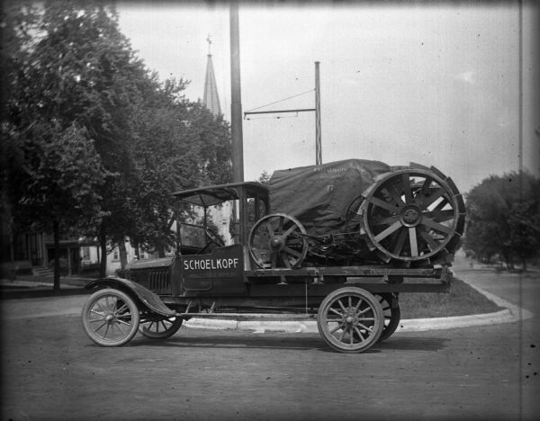 Schoelkopf truck with a Fordson tractor on the truck bed is parked outside the Schoelkopf Lincoln Ford Dealership at 210-216 E. Washington Avenue. The church steeple in the left background may be St. John Lutheran Church. Behind the truck is a railroad warning arterial.