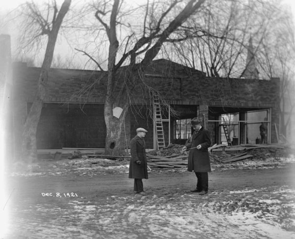 View from street towards construction of the Mazo Motor Company, which sold and serviced Ford and Fordson vehicles and agricultural machines. Standing in the street in the foreground are two unidentified men, one of whom is holding a measuring tape.