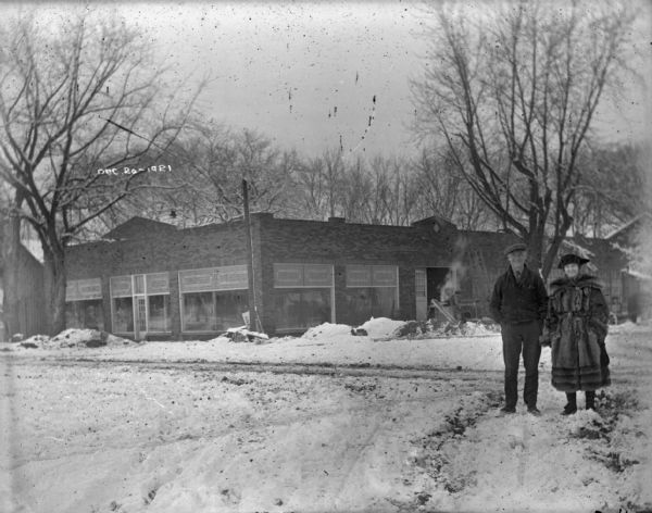 View from street of the construction of the Mazo Motor Company, which sold and serviced Ford and Fordson vehicles. Standing in the snow in the right foreground are a smiling man and woman, unidentified. The woman is wearing a large fur coat.