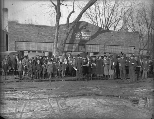 View across street towards a large crowd of people posing outside of the Mazo Motor Company dealership at the opening in early spring. Behind the crowd are parked automobiles. The sign on the building says: "Sales and Service, Ford-Fordson, Cars, Trucks and Tractors, Mazo Motor Company."