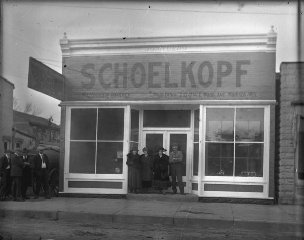 View from street of a group of three woman and one man standing in the doorway of the dealership in Mazomanie, where, in late 1919 or early 1920, Louis Schoelkopf opened a branch of his Ford dealership. Ford automobiles are displayed in the show windows. Standing on the sidewalk on the left are four men are standing in front of what may be a Fordson tractor. The words 'John Deno' are at the top of the building.