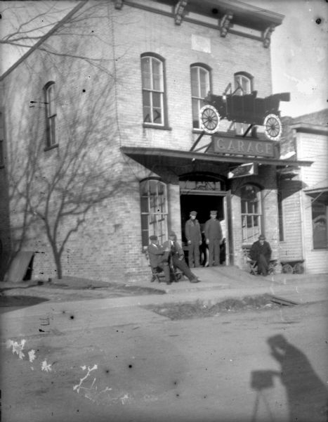View from street of five men posing outside a Ford garage, a two-story brick building in an unidentified small Wisconsin town. Above the entrance is a "Garage" sign below a small representation of a Ford Model T touring car. The shadow of the photographer is in the lower right foreground.