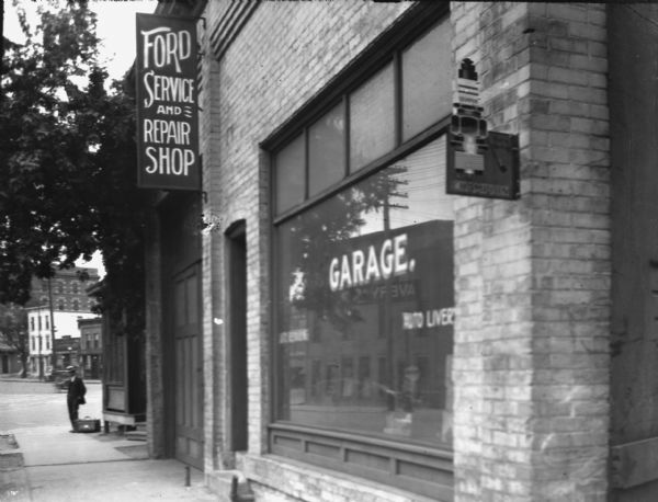 View along sidewalk of the front of the Ford Service and Repair Shop garage. This appears to be the block of Williamson St in the foreground. The Cardinal Hotel is visible in the background There is a Champion spark plug sign on the right corner of the shop. In the show window is a sign that reads: "Auto Livery." On the far corner, standing at the end of the sidewalk is a man, perhaps a salesmen with his sales kit.