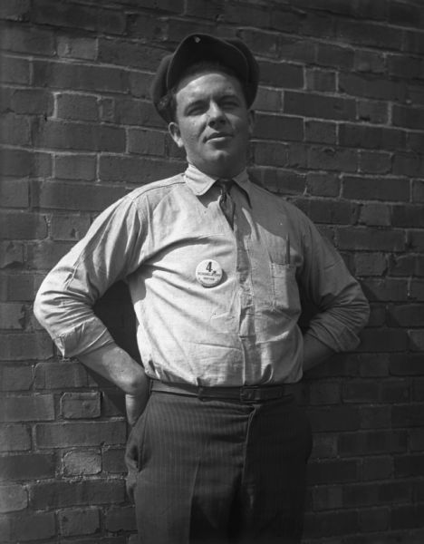 Portrait of a Schoelkopf Service superintendent posing with hands on hips against a brick wall outdoors. He has a button on his shirt that reads: "4, Schoelkopf Service Superintendent.