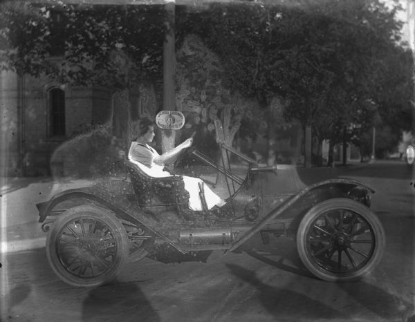 View from street towards Emma Schoelkopf who is driving a Ford automobile. On a tree behind her is a sign reading "School Grounds, Safety First, Autos Drive Slowly." In the foreground are the shadows of four men on the street.
