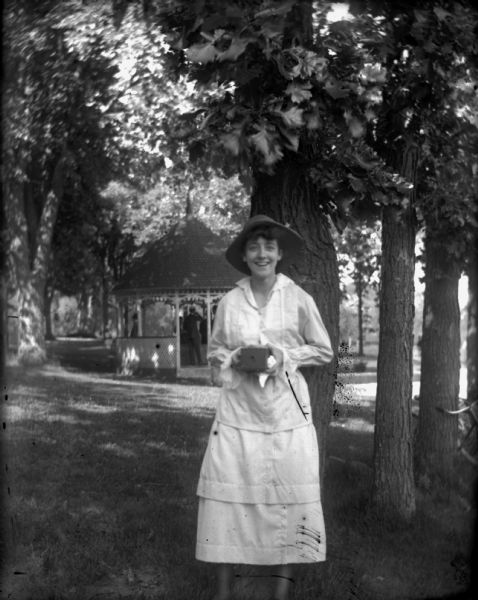 Emma Schoelkopf smiling and posing in a park with a camera in her hands. She is wearing a hat and a light-colored dress. In the background is a pavilion under trees.