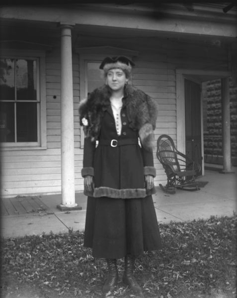 Emma Schoelkopf is posing in the yard in front of a house on Jenifer Street. She is wearing a matching skirt and fur-trimmed coat and hat. There is a rocking chair on the porch, and leaves are on the ground.