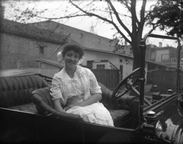 Emma Schoelkopf is sitting in the driver's seat of a Ford touring car. She is wearing a light-colored summer dress and smiling. Emma, the wife of Louis Schoelkopf, may have been the first female driver in Madison.