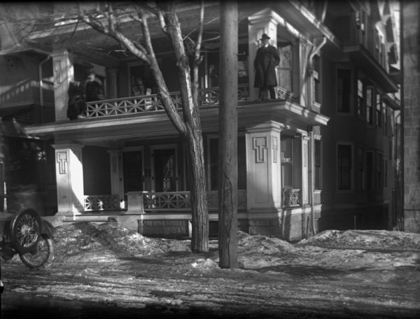 View from street towards three people standing on the second floor balcony of the Schoelkopf house at 137 Langdon St.  To the left you can see the edge of the Levi Vilas Mansion (no longer there).  The man on the right is standing just outside of the porch railing. Snow is on the ground. The house is at least three stories and has two flats.