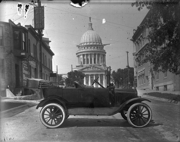 Ford Model T touring car parked on the street near the Wisconsin State Capitol building. In the background a banner strung above the street in front of the Capitol reads: "See Wisconsin Tractor Demonstration." The Wisconsin Tractor Demonstration was held in Madison in September 1916, one of eight held in midwestern states. Louis Schoelkopf sold Fordson tractors out of his dealerships.