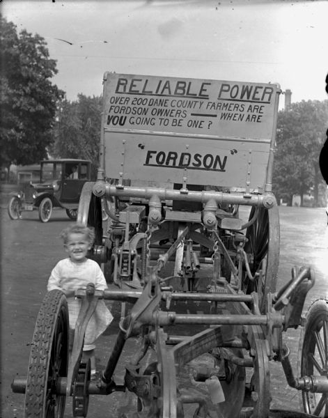 View of front of young girl standing at the side of a Fordson farm machinery. A sign on the tractor reads: "Reliable Power. Over 200 Dane County farmers are Fordson owners — When are you going to become one? 'Fordson'." There is man in a Ford pickup truck in the background.

Fordson tractors were manufactured by the Henry Ford Company from 1916 through the 1930s all over the world, but only sold from 1918-1928 in the United States.