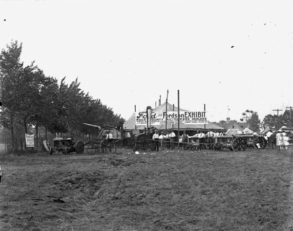 View across field towards a large canvas tent set up for the Ford and Fordson Exhibit by the Rock County Authorized Dealers. There are a number of Fordson tractors and other farm machinery arranged outside the tent, and groups of men are standing and posing around the tractors. On the right is a crowd of men, women and children. In the far background is a Ferris Wheel near a large building.

The authorized dealers are from Janesville, Beloit, Evansville, Milton Junction, Edgerton, Orfordville, and Clinton.

Fordson tractors were manufactured by the Henry Ford Company from 1916 through the 1930s all over the world, but only sold from 1918-1928 in the United States.