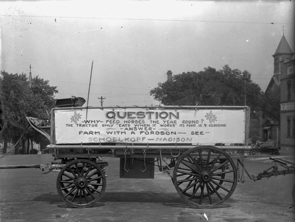 View down street towards a wagon with a sign on the side that reads: "Question: Why Feed Horses the Year Round? The Tractor Only "Eats When It Works". Its Food is 9 [cent] kerosine. Answer: Farm with a Fordson, See Schoelkopf — Madison."

The wagon is parked in front of the Schoelkopf Ford dealership at 212 E. Washington Avenue.