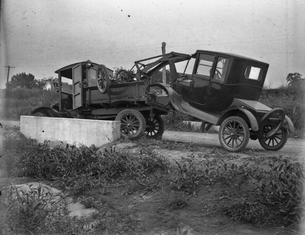 Left side view from roadside of a tow truck pulling a 1919 Ford coupe. The detached front axle of the coupe is in the bed of the tow truck.