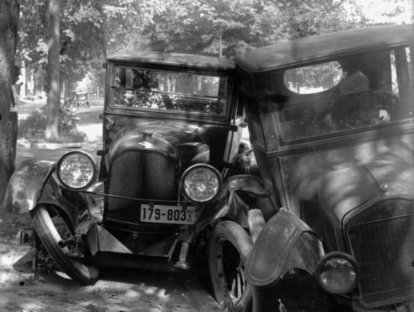 View of the front of two automobiles, one of them a Ford Model T (right),  with crumpled fenders resting against each other near the curb. The car on the left also has a broken or bent axle with a license plate that reads: "179-803 Wis 21."

