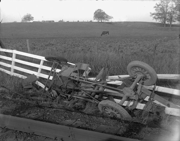 View over railroad tracks towards a train utility vehicle lying upside down against a broken fence. In the background is a cow in a field, and farm buildings are on the horizon in the far background.