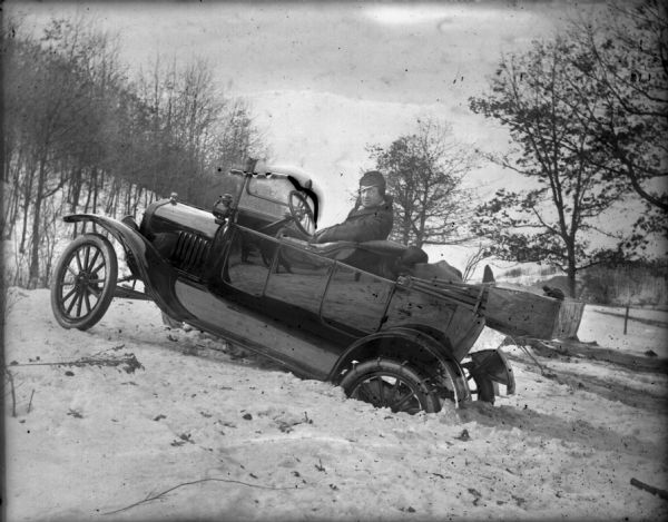A man sits in a Ford Model T touring car which is stuck in the snow on a hill in a rural area.