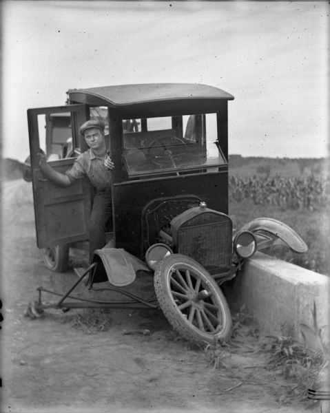View towards a man posing in the open door of an early Ford automobile, which has run off the road up onto a concrete barrier. The axle is broken and one tire is missing.