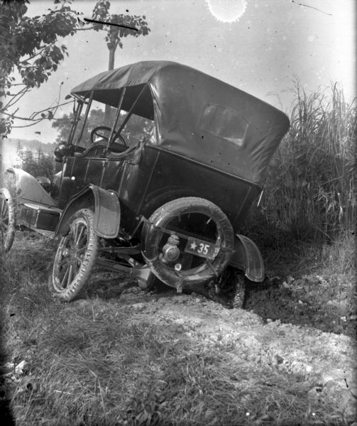 Three-quarter rear view from left of a Ford touring car which has run off the road into a ditch. It appears to be stuck in the mud. The license plate, which is broken on the right side, has a single star with the number 35.