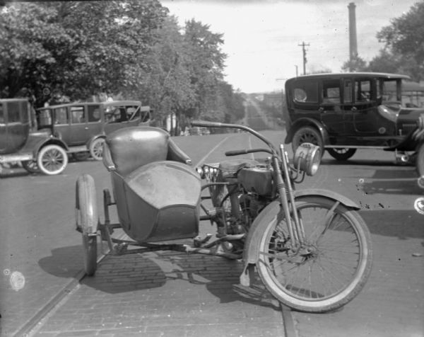 View down East Washington Avenue towards a motorcycle with a sidecar. It is parked outside the Schoelkopf Automobile dealership (not shown). The motorcycle has a large headlight and fenders on the tires. In the background are a number of Ford automobiles.