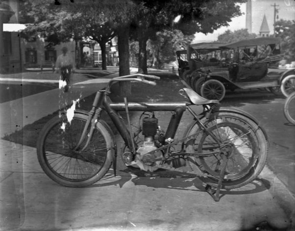 Side view of a motorcycle parked in a driveway along East Washington Avenue, near Schoelkopf Automobile Dealership (not shown). There are several automobiles parked along the curb in the background. A man is walking up the sidewalk on the left.  The motorcycle may be a Pierce.