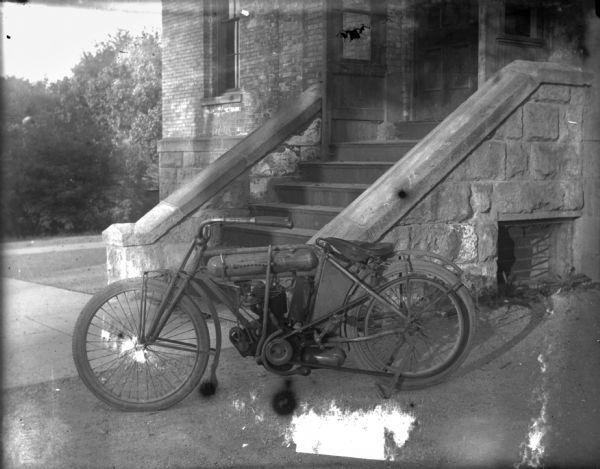 Left side view of a motorcycle parked near the steps of a building. The motorcycle may be an M-M (Marsh-Metz) or a clone by Peerless or Haveford.