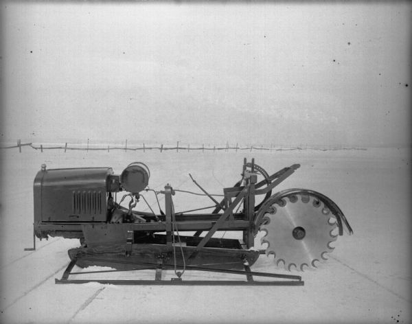 Left side view of a brand new ice harvesting machine sitting on a frozen lake covered with snow. The machine is sitting over tracks and cuts in the ice, probably made by the machine. In the background is a wood fence on the lake.