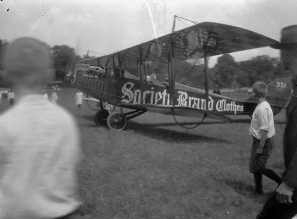 Left side view of a man driving the Corben Jenny with "Society Brand Clothes" painted on the fuselage. The airplane is moving down a field. In the foreground are several men and boys walking along with the airplane. In the background are more people approaching the airplane.