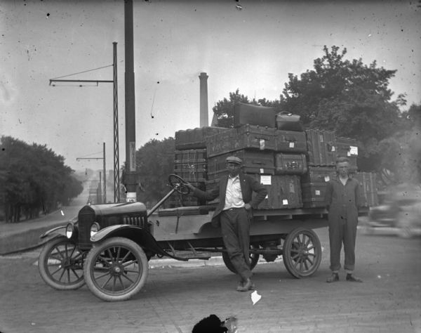 Two men pose standing beside a Ford delivery truck piled high with baggage. The truck is parked near the top of East Washington Avenue.