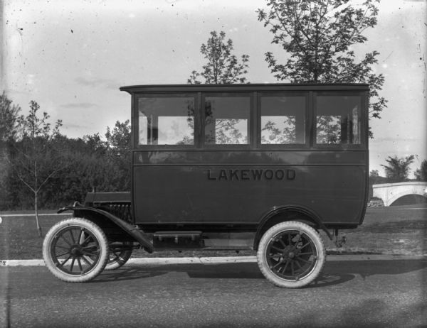 Left side view of a Ford school bus, with the word "Lakewood" painted on the side, parked on a street. In the background are trees and a bridge.