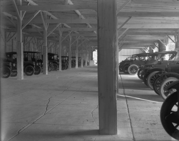 View of automobiles in a parking garage. They are parked in rows on the left and right. A man is standing in the far background at the end of the garage.